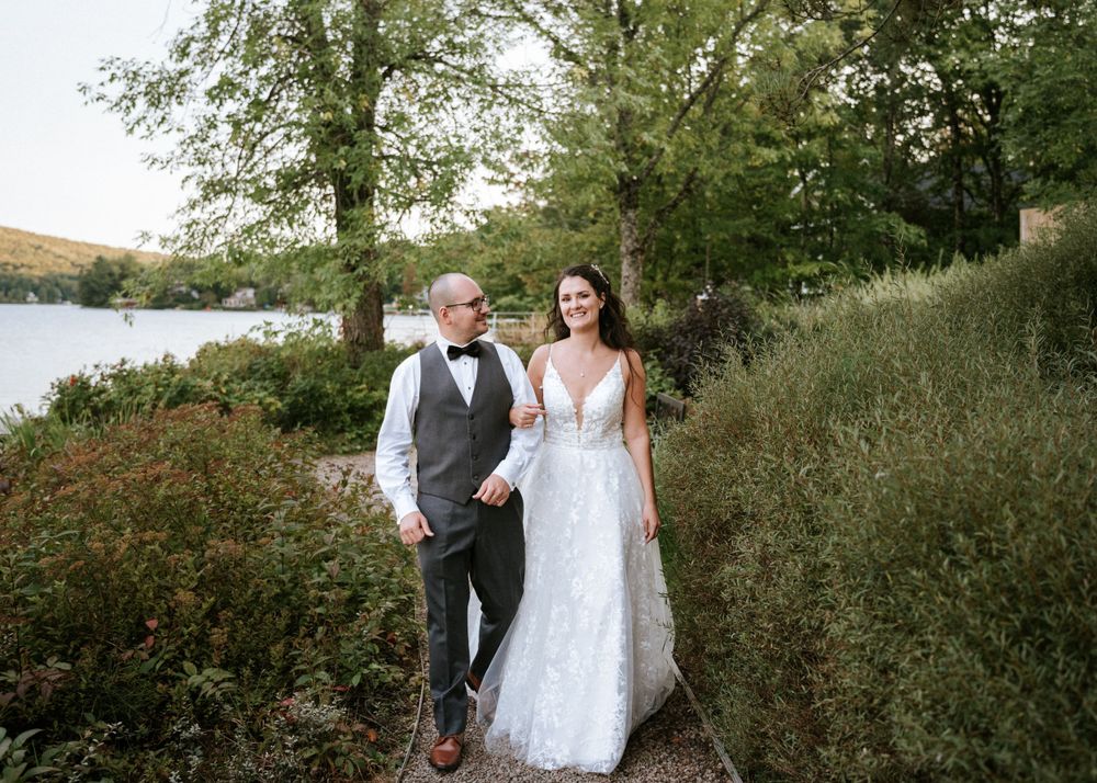 Un couple en tenue de mariage se promène le long d'un sentier pittoresque au bord d'un lac, entouré d'une végétation luxuriante.