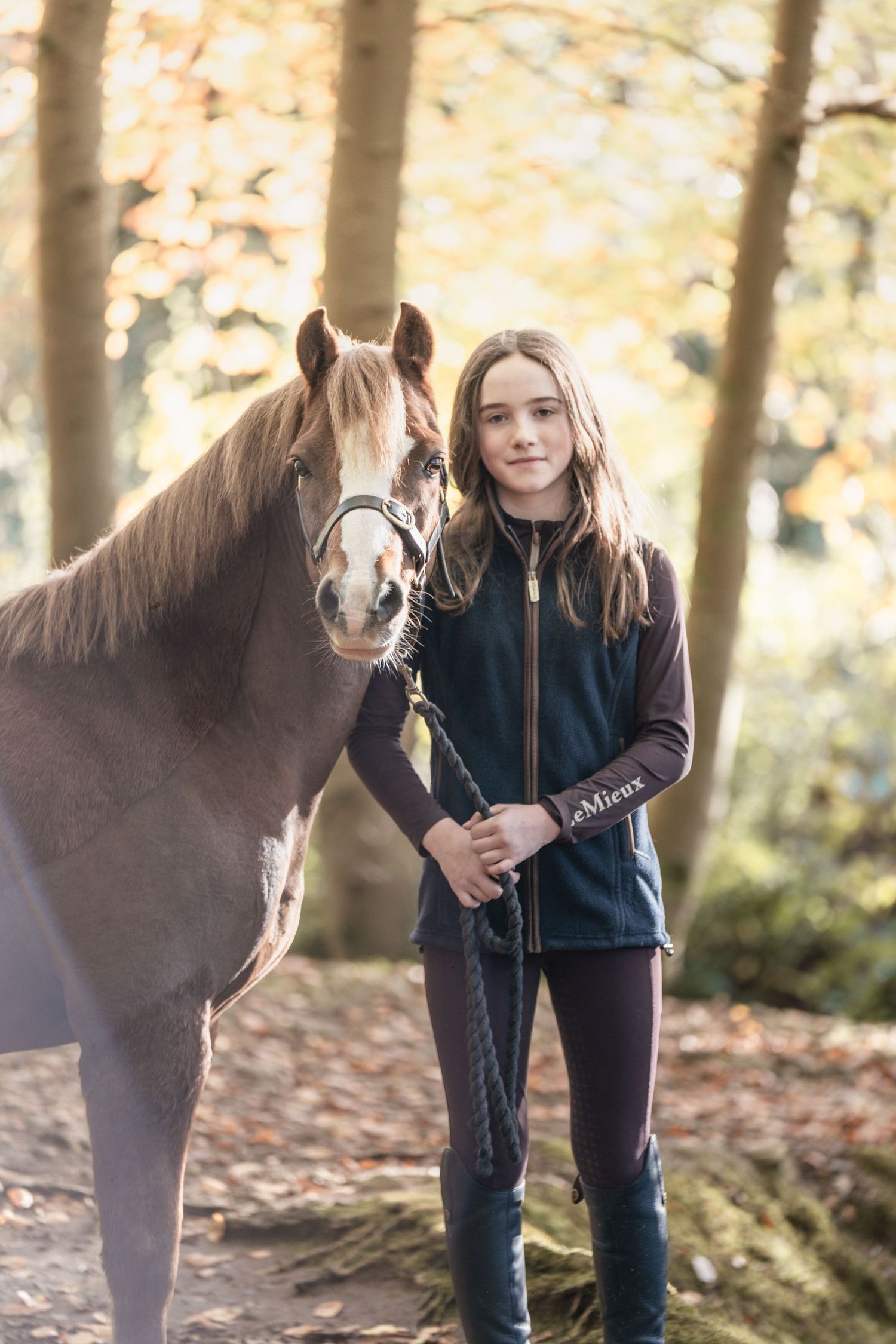 A brown pony stands with its handler in a wooded area with autumn leaves scattered on the ground.