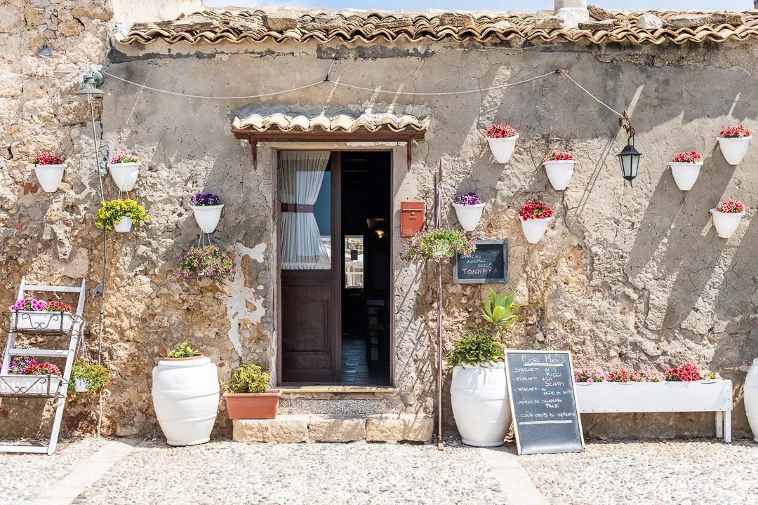 A rustic stone shop entrance decorated with hanging pottery and plants sits along a sunny street.