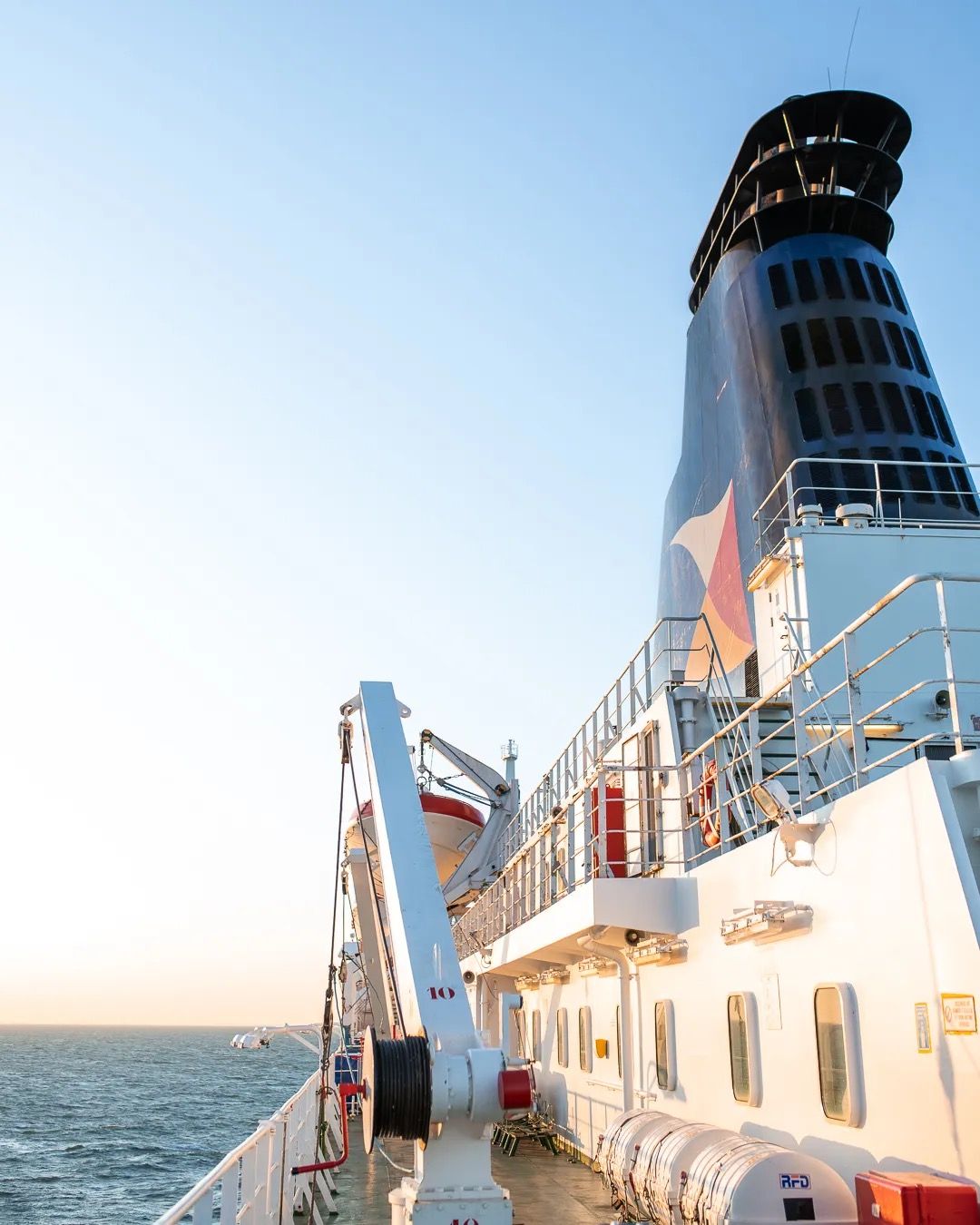 View of a ferry deck and funnel sailing across calm sea in bright daylight.