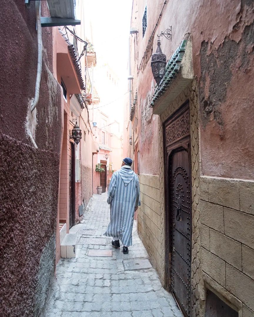A person walking through a narrow, sunlit alleyway between tall terracotta buildings.