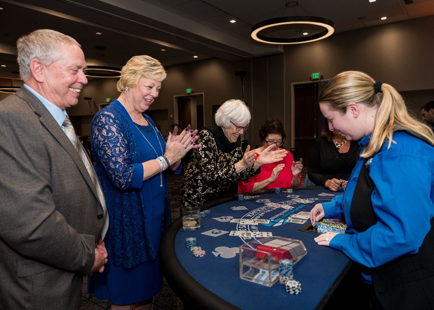 People gather around a casino gaming table with blue felt while a dealer in blue interacts with guests.