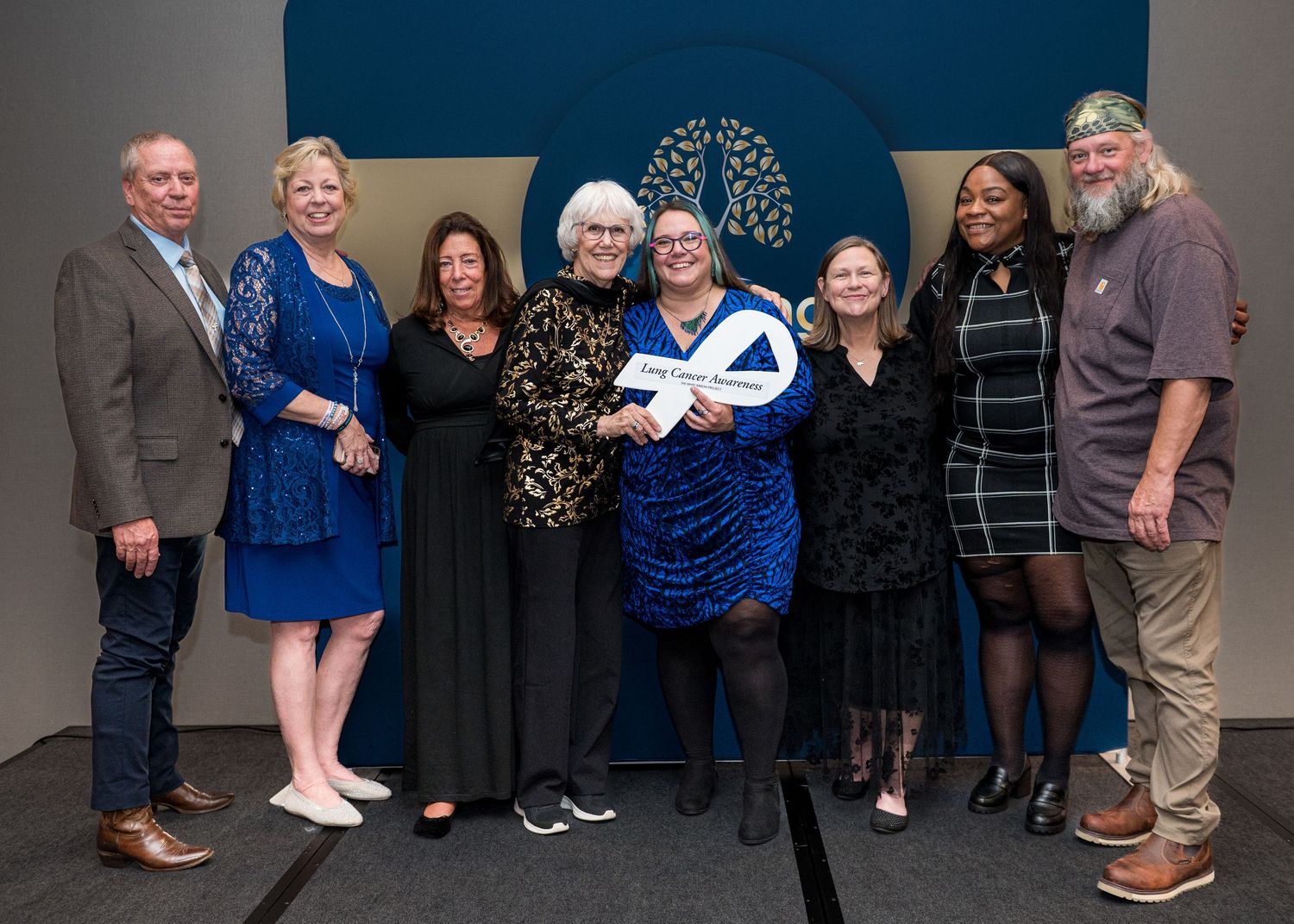 A group poses together against a blue backdrop with a decorative tree logo.