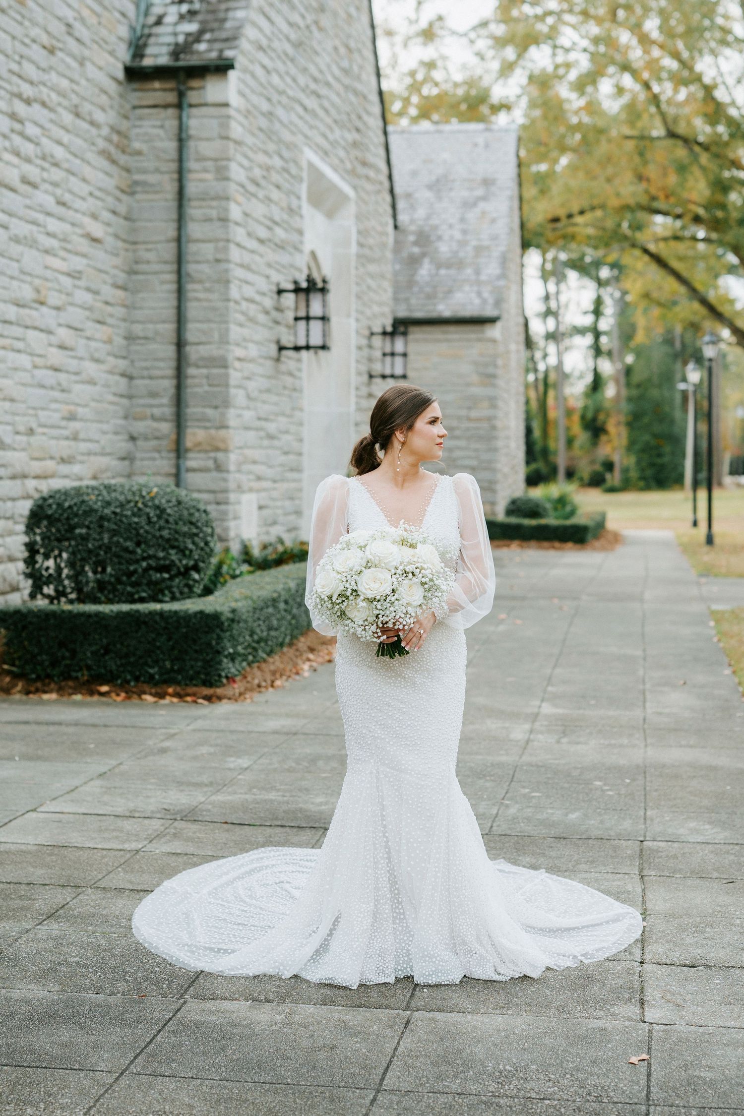 Elegant bride in long-sleeved lace wedding gown holds white bouquet while standing on stone path outside church.