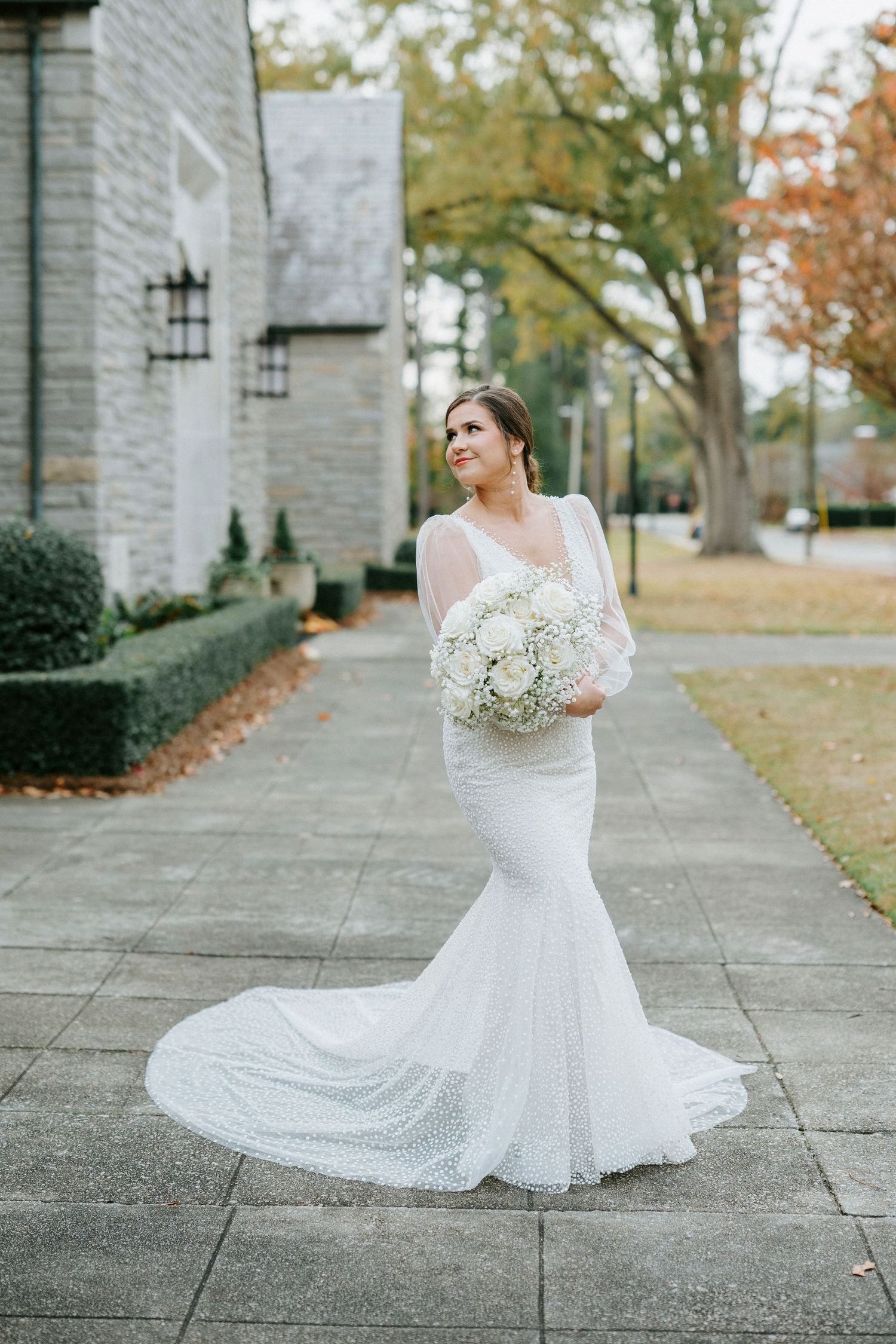 Elegant bride in fitted lace mermaid wedding gown holds white bouquet on stone path during autumn wedding.