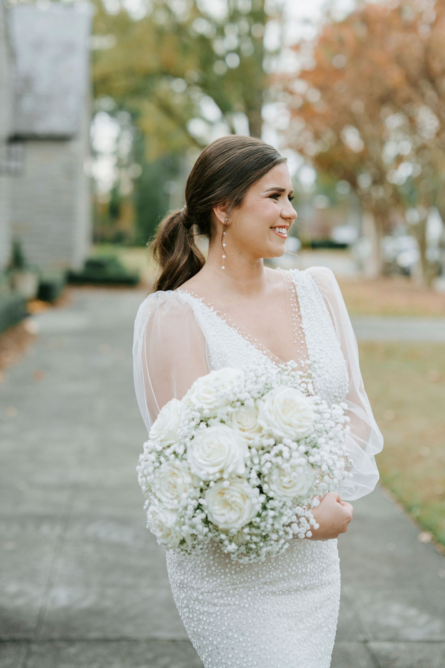 A bride in a fitted lace gown with sheer sleeves holds an elegant white rose and baby's breath bouquet outdoors in autumn.
