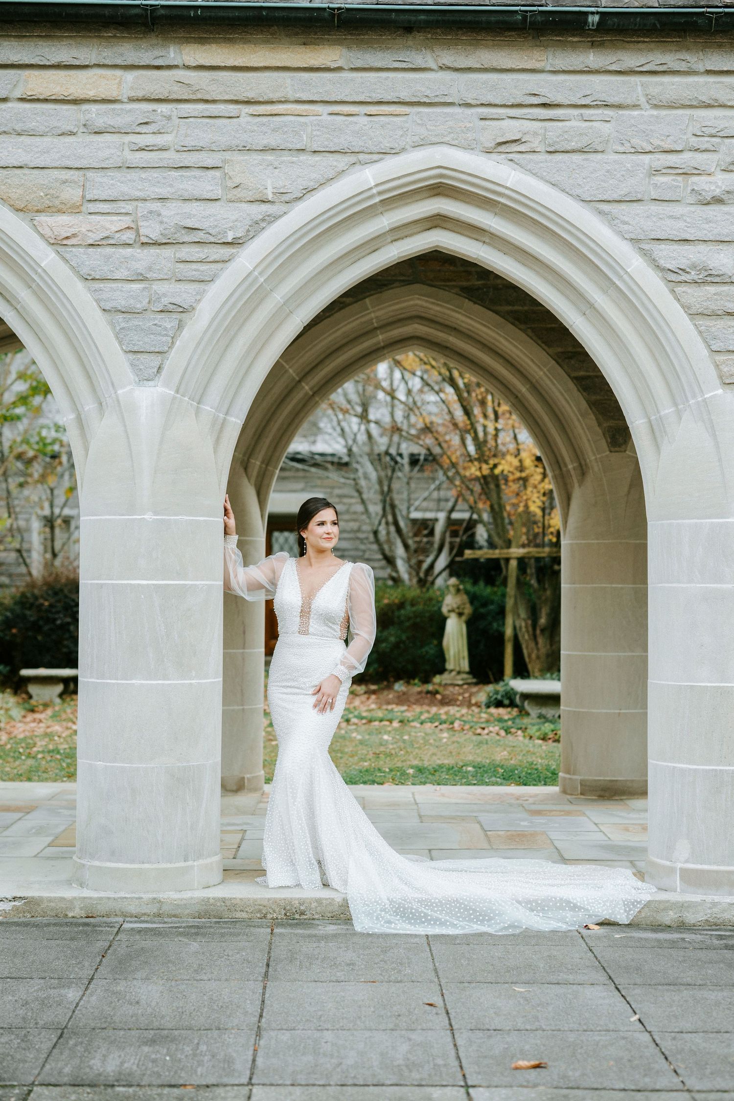 A stunning wedding gown with a mermaid silhouette and long sleeves photographed under a gothic stone archway.