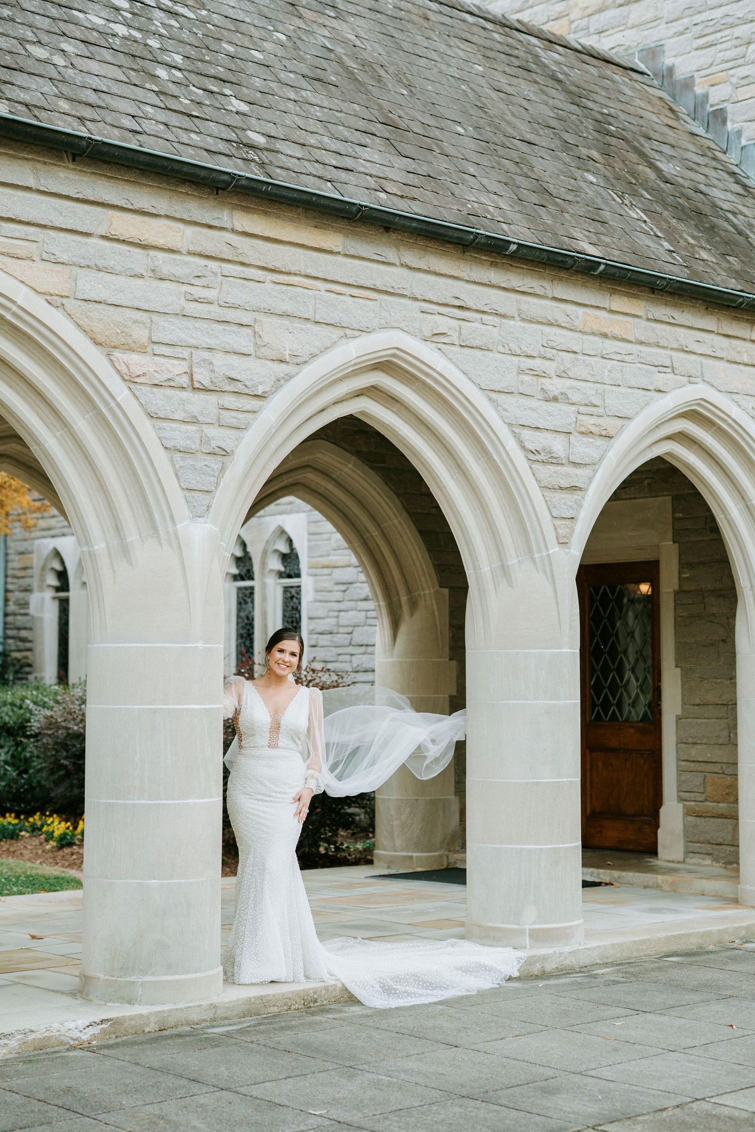 A flowing white wedding veil creates a dramatic movement effect under stone archways at a church or cathedral.