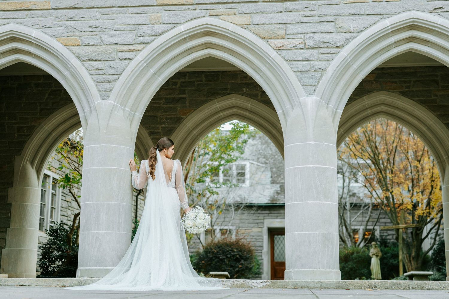A bride in a white wedding gown stands elegantly under a series of white stone archways in a garden setting.