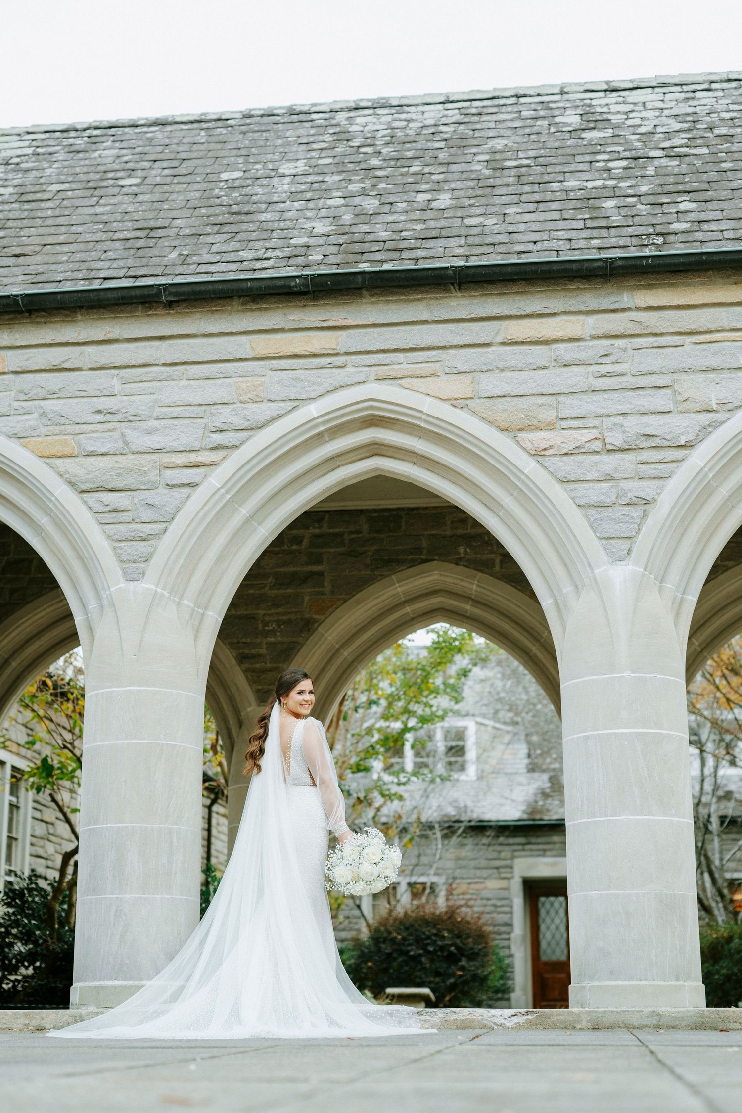 A classic bridal portrait captured from behind showing a white wedding gown with cathedral train under stone archways.