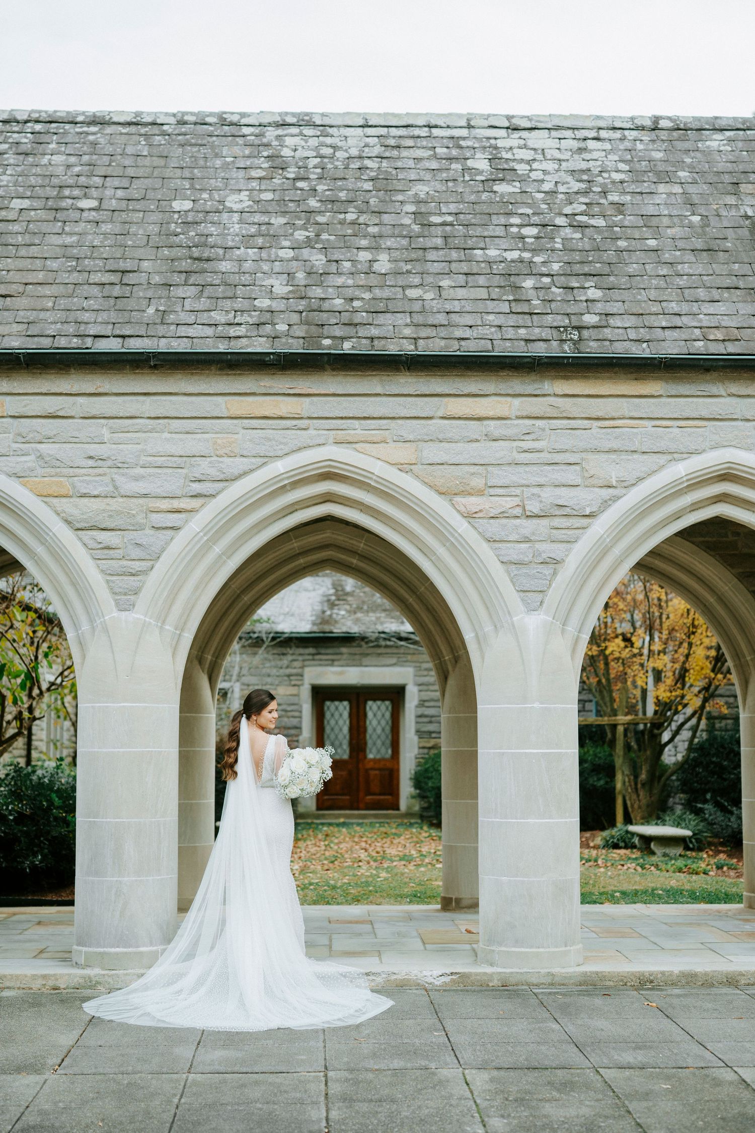 A bride in a flowing white dress stands beneath Gothic stone arches at a historic church entrance.