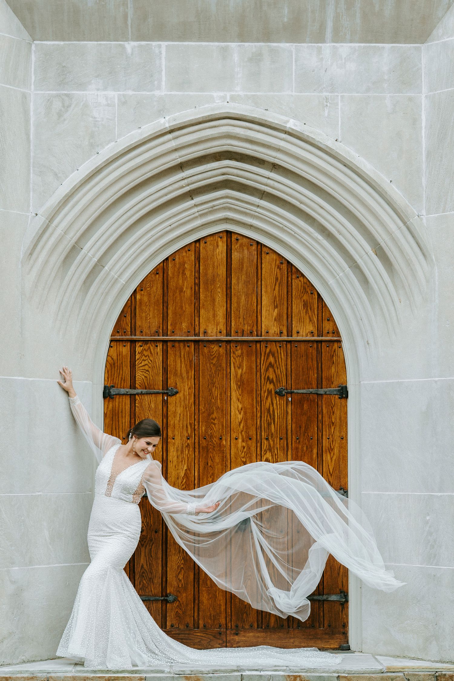 A flowing white veil catches the wind against a wooden church door with Gothic stone archway.