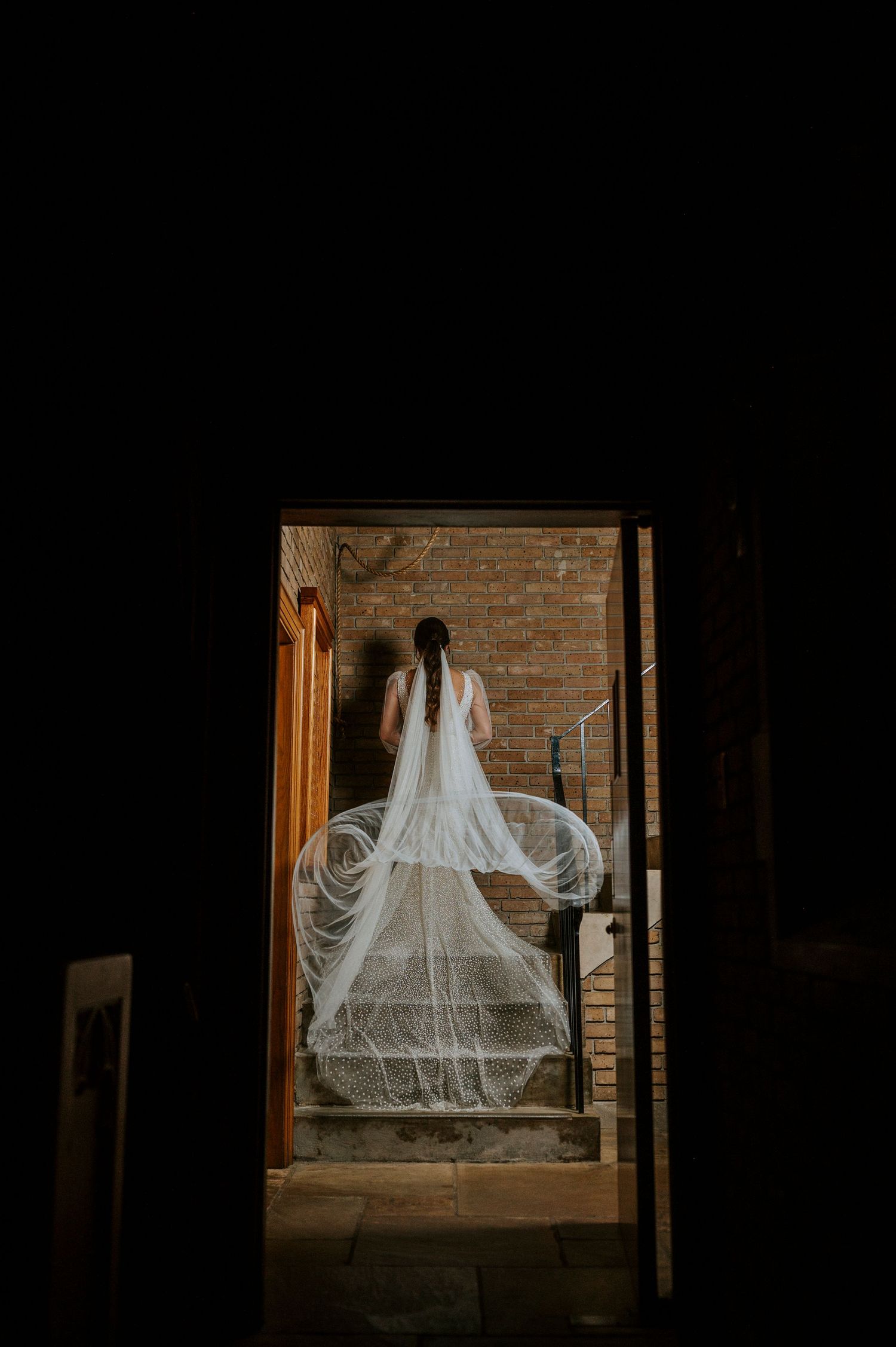 A wedding dress with a flowing veil is silhouetted in a rustic doorway against exposed brick walls.