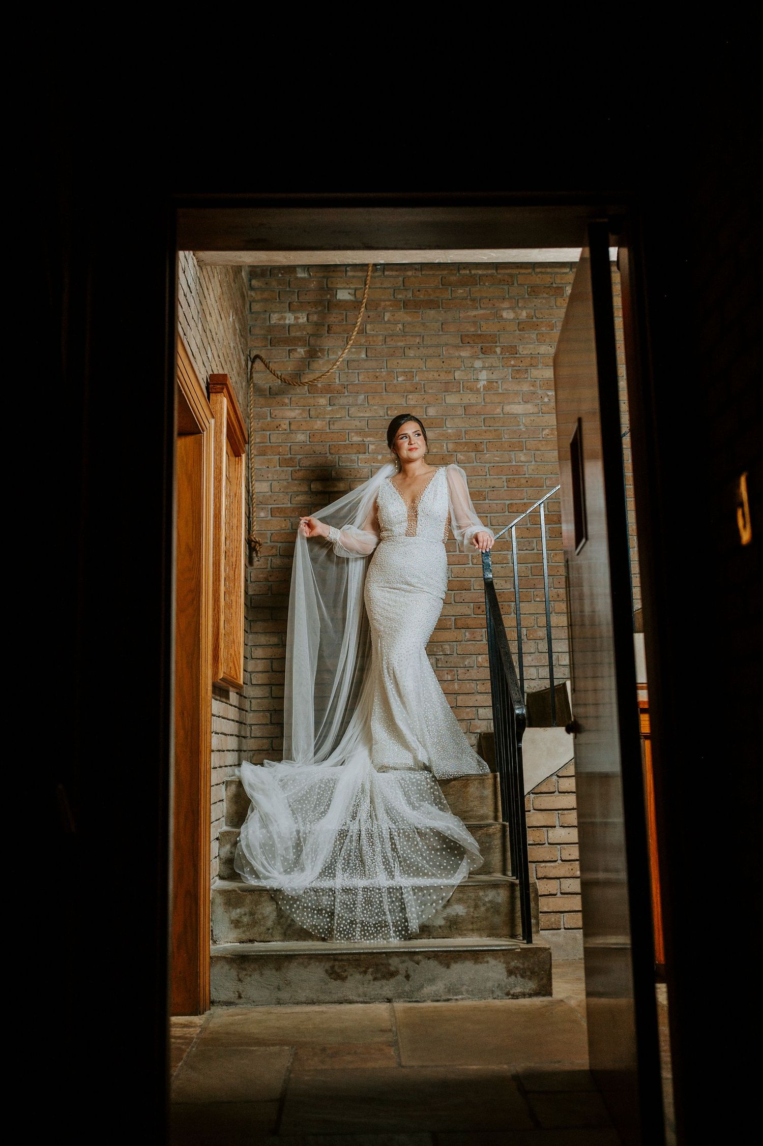 A long white dress and veil create an elegant silhouette against exposed brick walls and wooden doorframe.