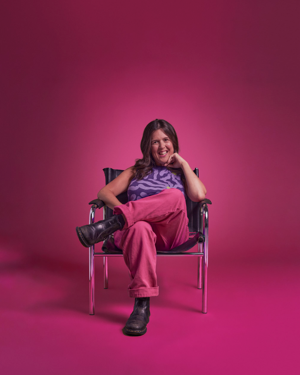 Person lounging and posing playfully on chairs against pink studio backdrop.