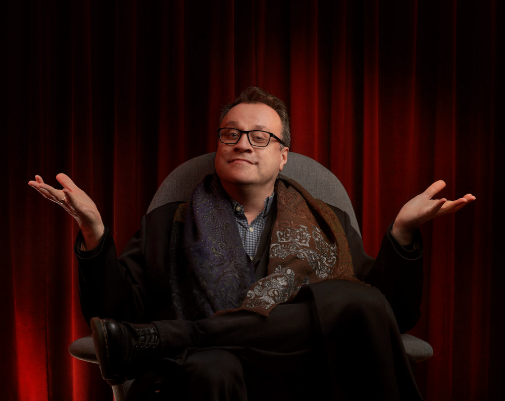 Person gesturing expressively while seated against red curtain background.