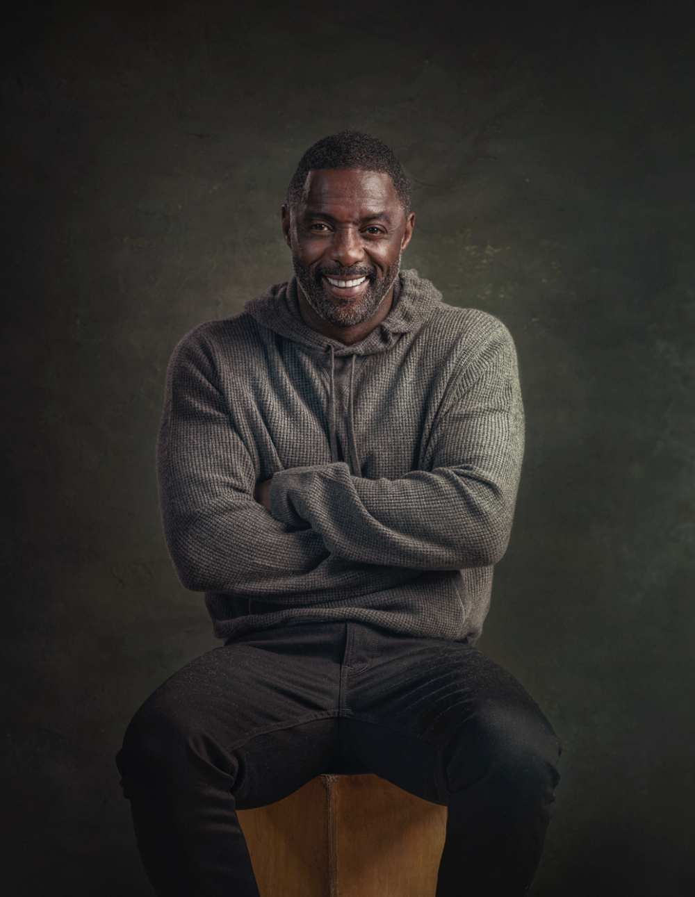 Person in grey sweater sits casually on chair against dark background in artistic portrait series.