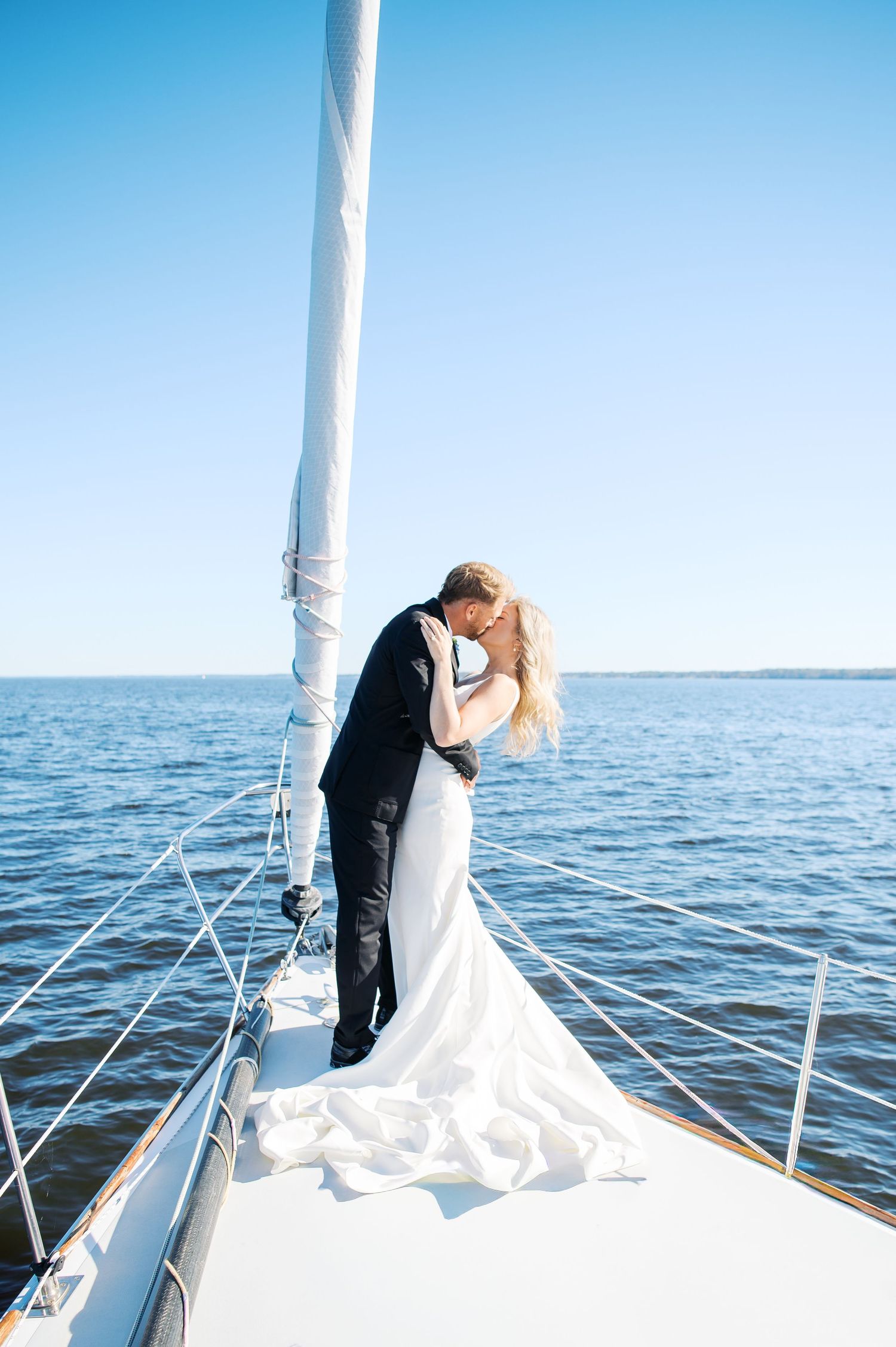 A romantic wedding couple embraces on the bow of a sailboat against blue ocean waters and clear sky. Photo by Heather Moss Photo