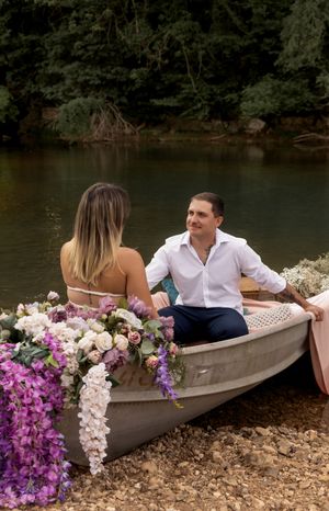 Decorated canoe with pink and purple flowers sits along shoreline as couple poses inside.