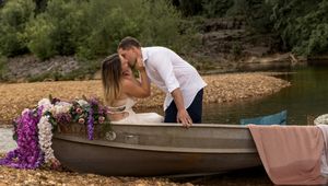 Couple shares intimate moment in flower-adorned boat against wooded shoreline.
