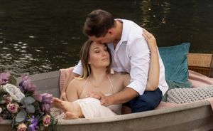 Romantic couple poses in rowboat decorated with purple and white flowers on tranquil lake at sunset.