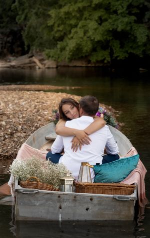 A romantic couple embraces in a decorated rowboat on a peaceful river with floral arrangements and pillows.