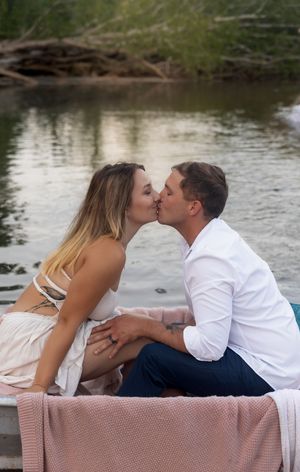 Romantic couple shares intimate moment while sitting on pink blanket by serene lake.