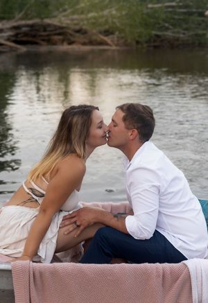 Couple embraces tenderly on pink blanket beside peaceful waterfront.