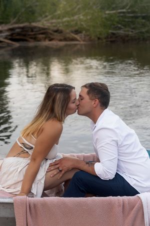 Loving couple shares romantic kiss on blanket near tranquil water.