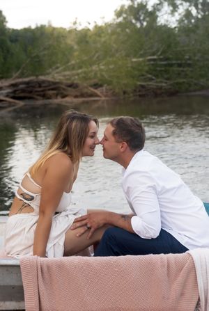 Couple in white and blue outfits share tender moment by lakeside.