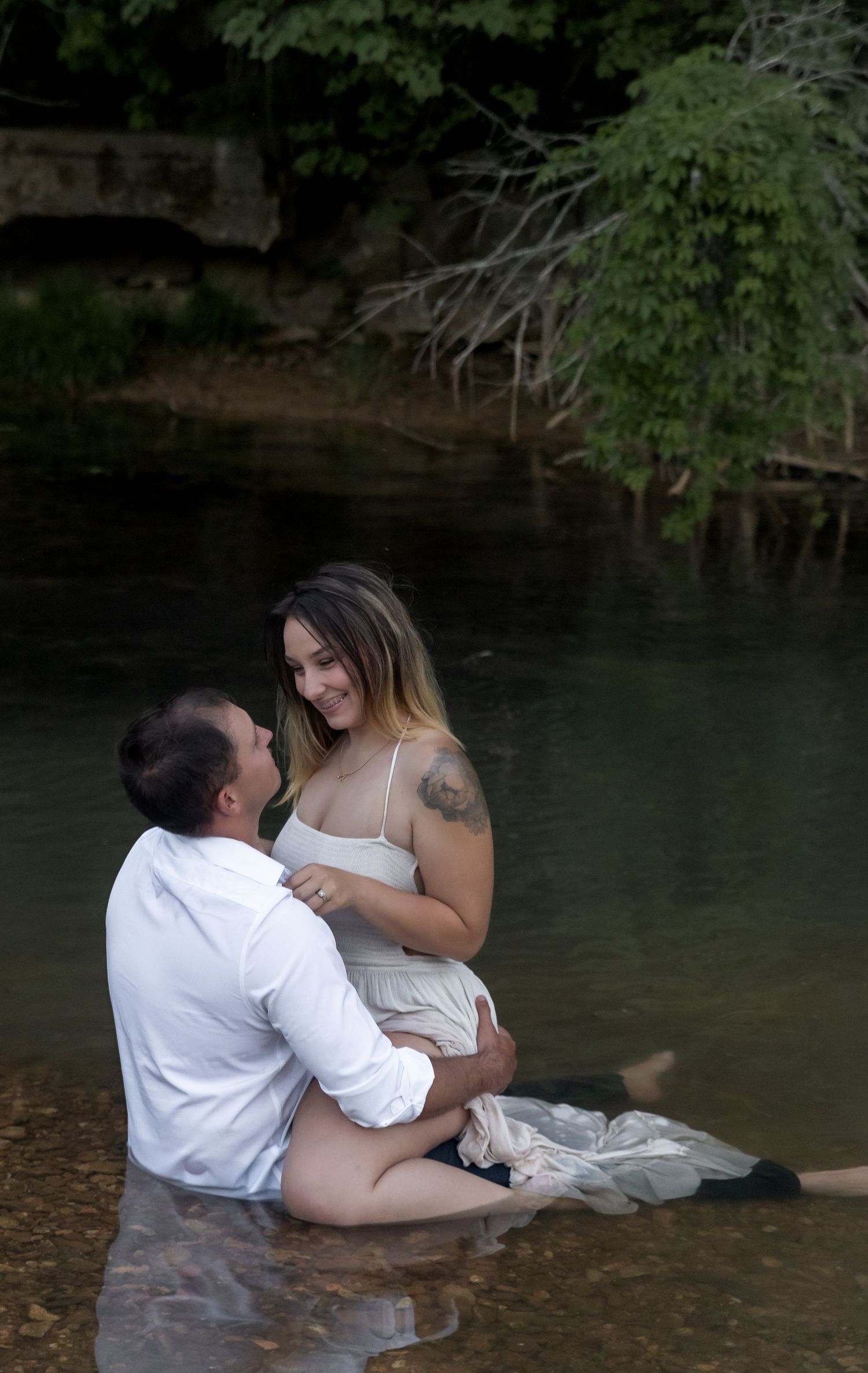 A couple embraces while sitting together in a tranquil stream during a photo session.