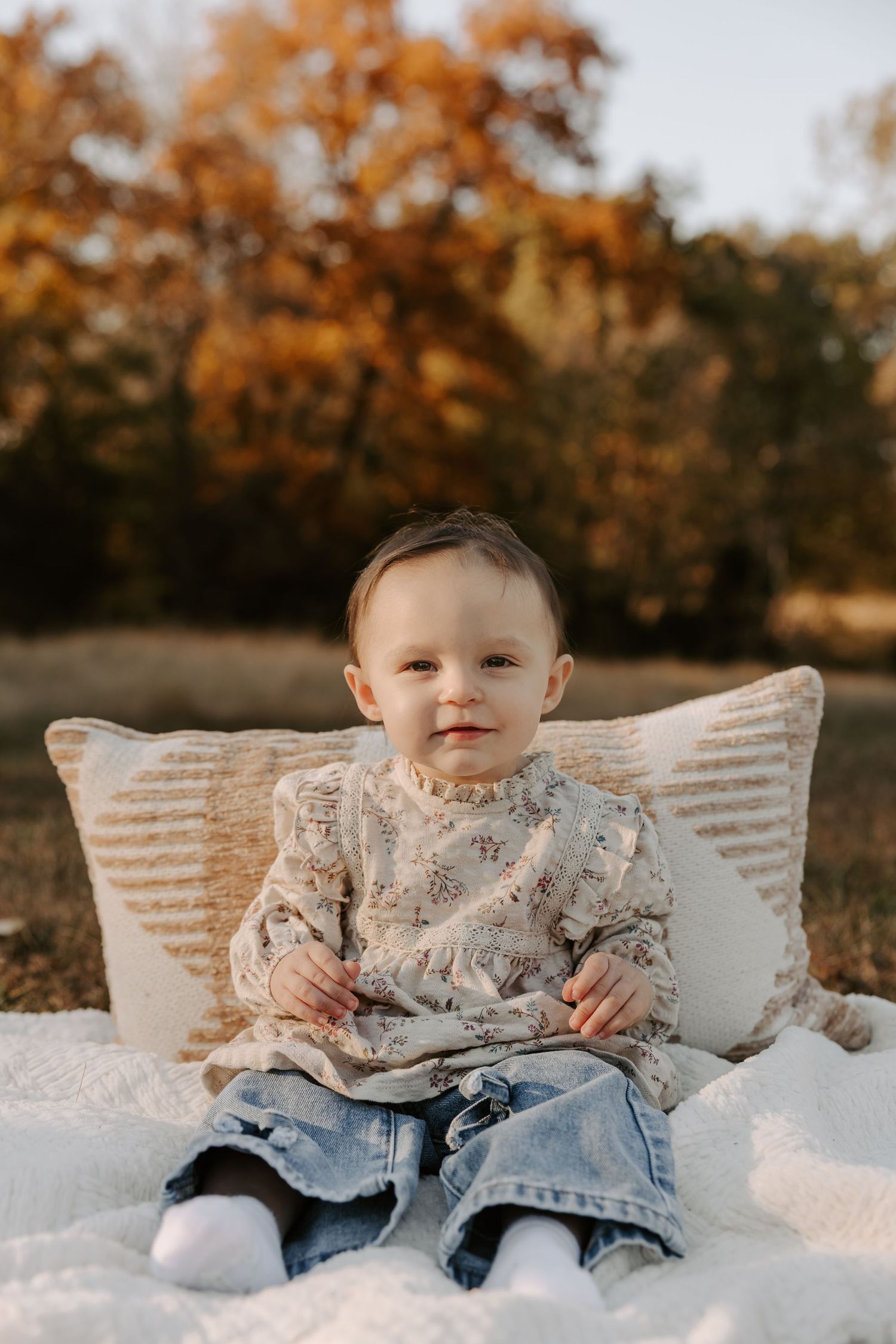 A toddler sits on white blankets and pillows outdoors during autumn wearing jeans and a patterned shirt.