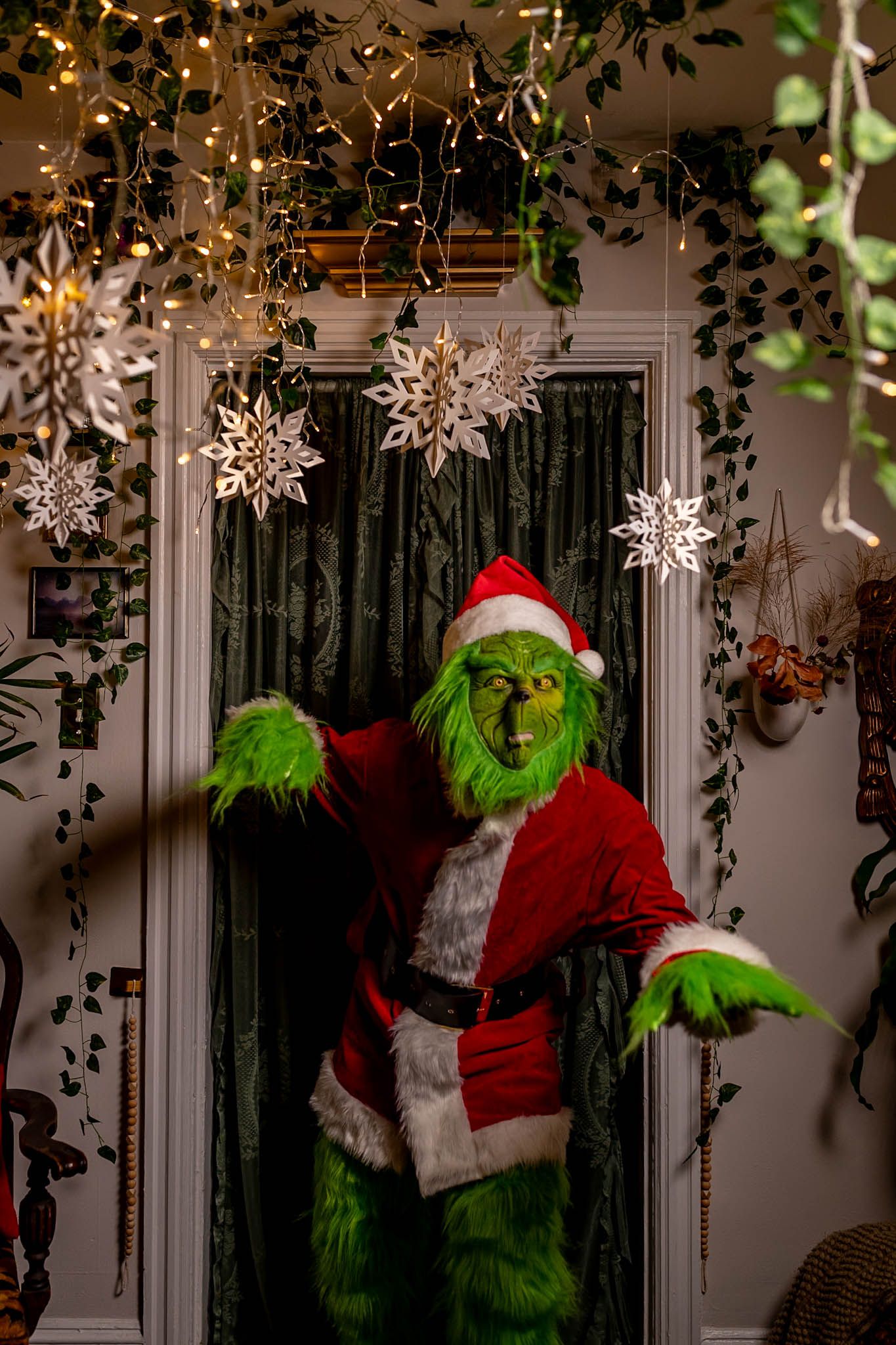The Grinch in a Santa suit poses dramatically against a backdrop of hanging snowflakes and greenery.