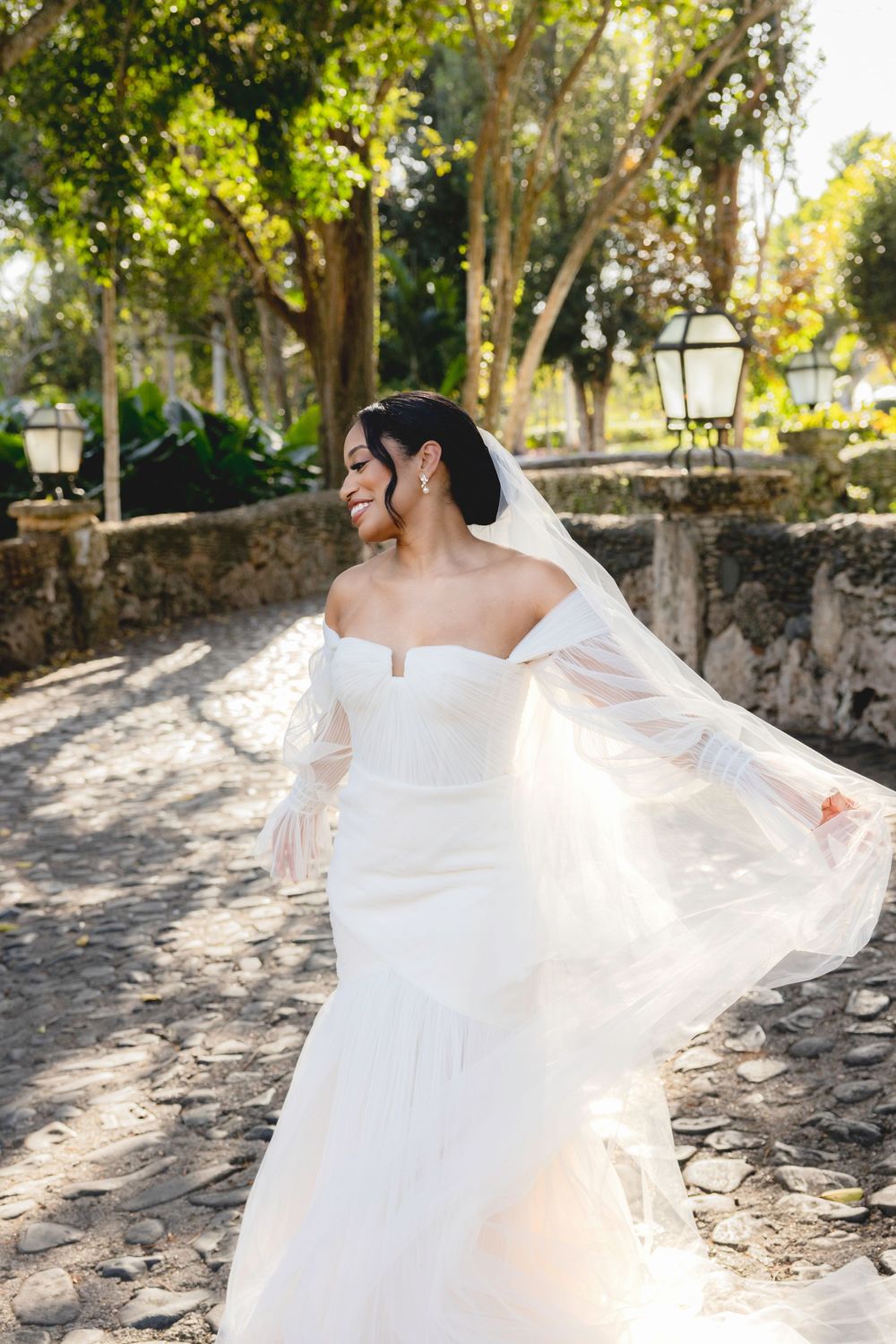 A bride in a flowing white wedding dress twirls gracefully on a sunlit stone path surrounded by trees.
