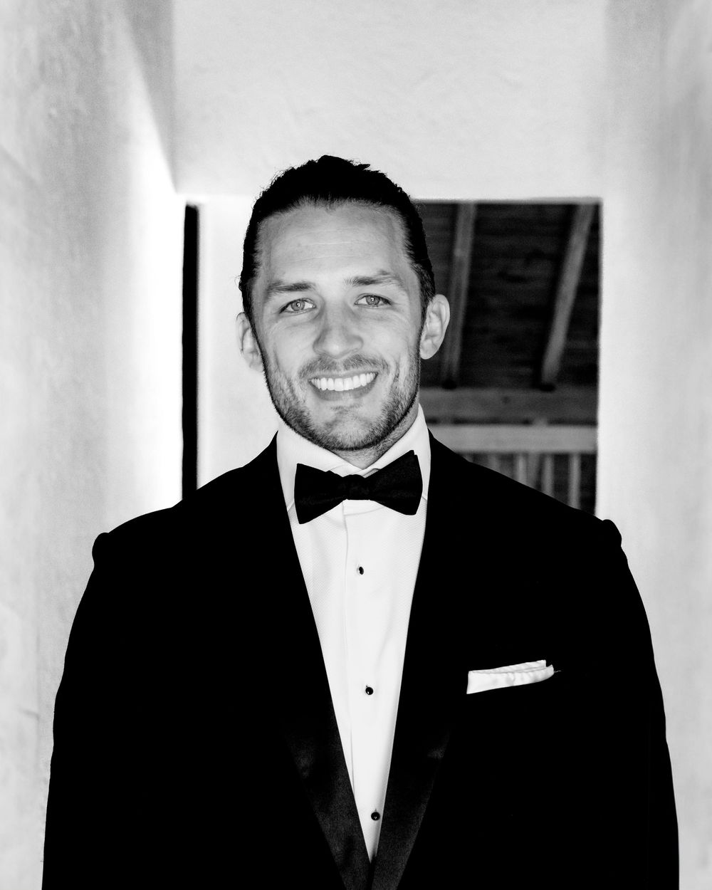 A black and white portrait of someone in a classic tuxedo with bow tie and white pocket square, smiling warmly.
