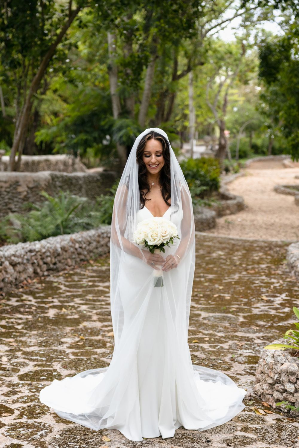 A beautiful bride in a flowing white wedding gown poses outdoors on a natural path surrounded by trees.
