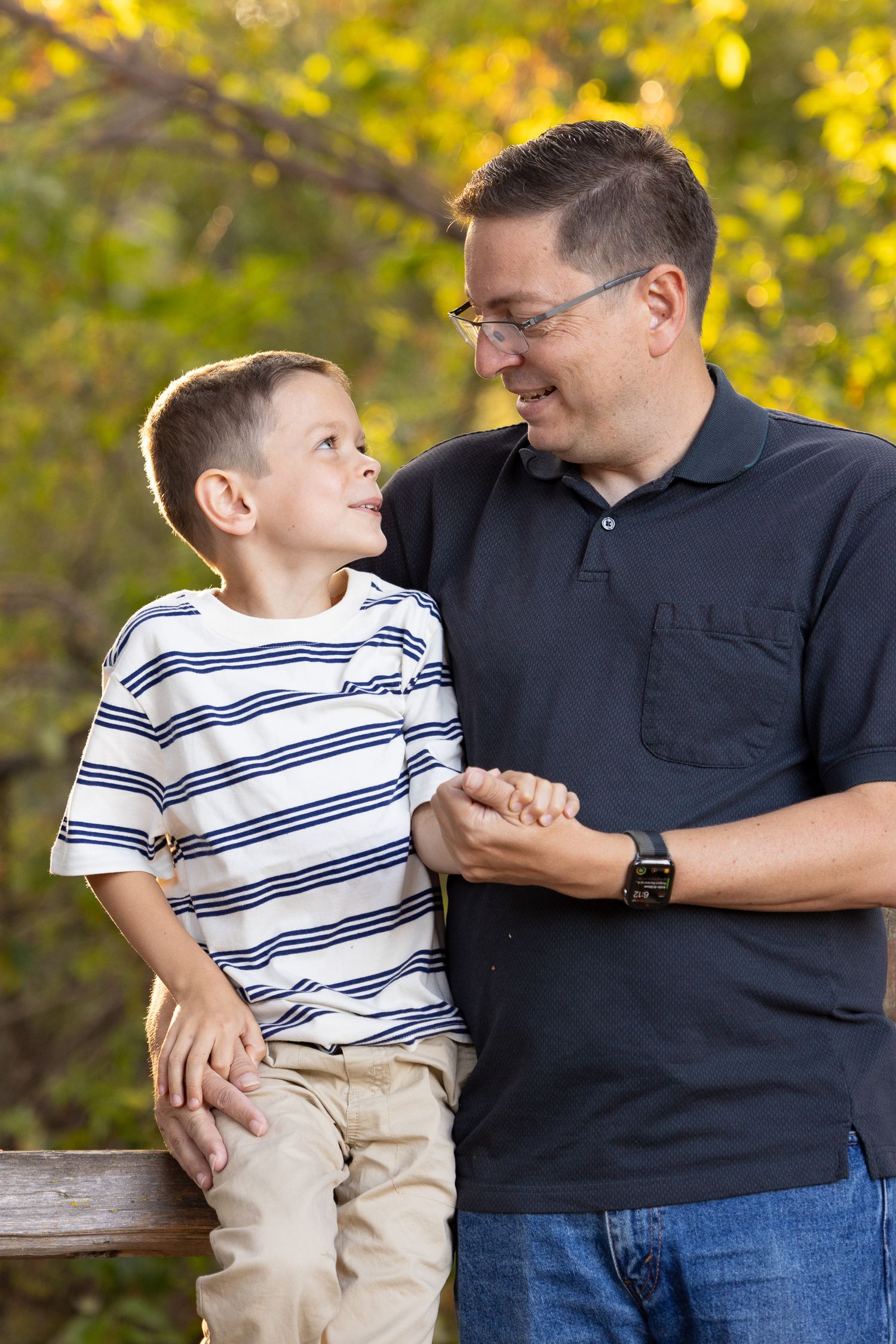 A father and young son smile at each other against an autumn backdrop during golden hour.