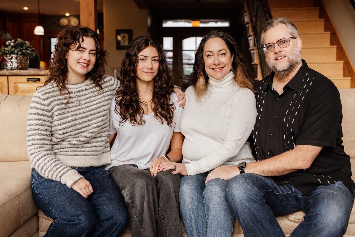 A family sits together on a brown sofa in coordinated casual outfits for a family portrait session in their home.