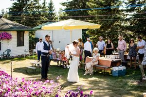 An outdoor wedding reception setup with pink flowers and guests gathering under trees.