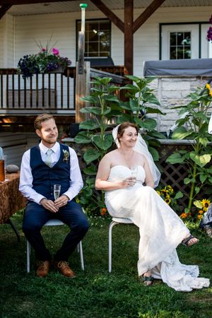 Couple in wedding attire sits together on white chairs in a garden setting.