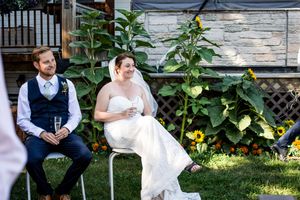Wedding couple sits together on white bench in garden surrounded by lush greenery and yellow flowers.