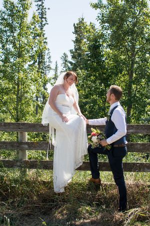 Playful wedding couple poses by wooden fence in natural outdoor setting.