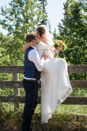 A young child in formal wear stands next to a flowing white wedding dress against a rustic wooden fence.