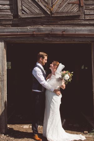 A newlywed couple embraces in front of a weathered barn entrance while holding a floral bouquet.