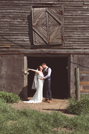 A bride and groom take creative photos along a weathered wooden barn wall.