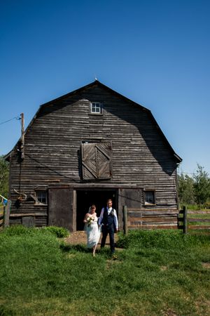 A rustic wooden barn with weathered siding stands against a bright blue sky on a grassy hillside.