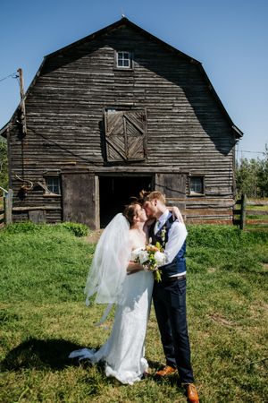 A rustic barn wedding photoshoot with a couple in white dress and navy suit against weathered wooden buildings.