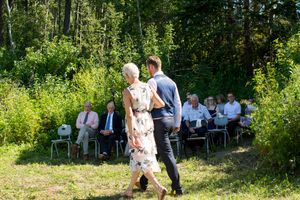 Outdoor wedding ceremony in a lush green forest setting with guests seated on wooden benches in the sunlight.
