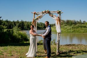 A wedding ceremony takes place under a wooden arch by a serene lakeside.