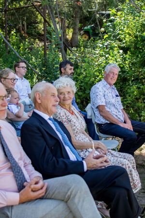 Guests seated outdoors at a garden event wearing formal attire and enjoying refreshments in the afternoon sunlight.